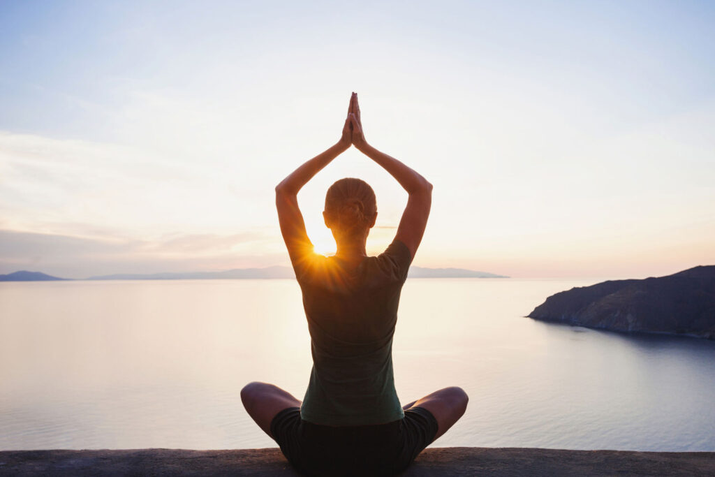 Frau in Yogapose blickt Richtung Sonnenuntergang